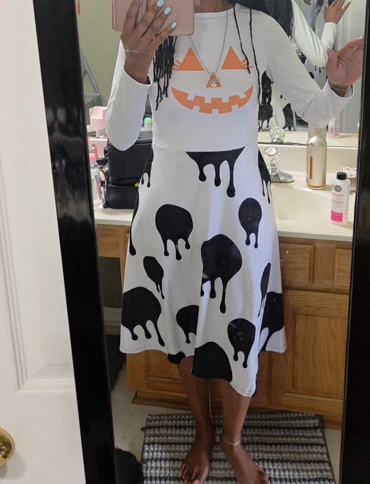 A person taking a mirror selfie in a bathroom wearing a white long-sleeve dress with an orange jack-o'-lantern face on the chest and black dripping shapes scattered across the skirt.
